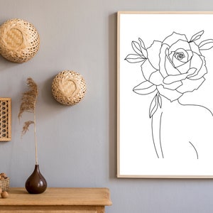 May include: A framed line art print of a woman's silhouette with a rose for a head. The artwork is in a light wood frame. Decorative woven wall hangings and a wooden table with a vase and decorative items are also in the image.