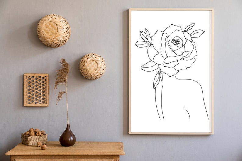May include: A framed line art print of a woman's silhouette with a rose for a head. The artwork is in a light wood frame. Decorative woven wall hangings and a wooden table with a vase and decorative items are also in the image.