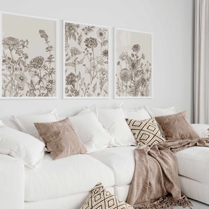 May include: Three framed botanical art prints with detailed line drawings of various flowers and plants in shades of brown and beige. The prints are displayed above a white sectional sofa with decorative pillows and a throw blanket.