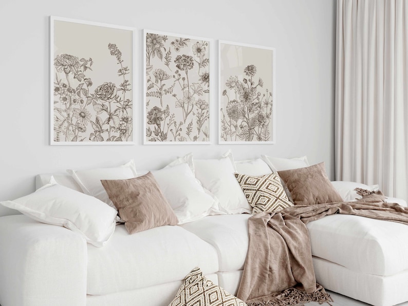 May include: Three framed botanical art prints with detailed line drawings of various flowers and plants in shades of brown and beige. The prints are displayed above a white sectional sofa with decorative pillows and a throw blanket.