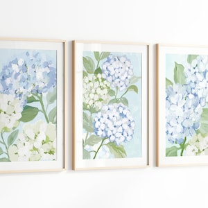 Pode incluir: Um conjunto de três impressões de arte de parede emolduradas com flores de hortênsia pintadas em tons de azul claro, branco e verde suave. Cada impressão retangular está inserida numa moldura de madeira clara, exibindo cachos de flores.