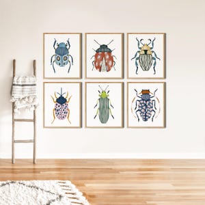 May include: Six framed insect art prints. Each print features a different colorful beetle illustration in shades of blue, red, green, and pink. The prints are displayed on a white wall above a wooden floor, with a wooden ladder and a rug in the foreground.