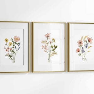 May include: Three framed floral art prints. Each print features a watercolor bouquet of flowers in shades of pink, yellow, and blue. The frames are gold-colored, and the background is white.
