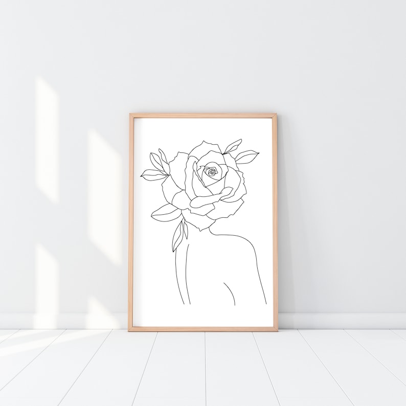 May include: A framed art print featuring a minimalist line drawing of a person with a rose for a head. The artwork is in black and white, with a simple, elegant style. The frame is a light wood color, and the background is white.