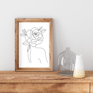 May include: A framed art print featuring a minimalist line drawing of a figure with a rose for a head. The artwork is in a wooden frame and is displayed on a wooden surface. Decorative vases are also on the surface.