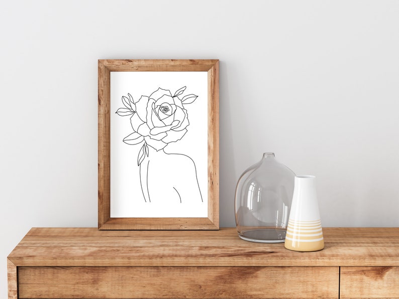 May include: A framed art print featuring a minimalist line drawing of a figure with a rose for a head. The artwork is in a wooden frame and is displayed on a wooden surface. Decorative vases are also on the surface.
