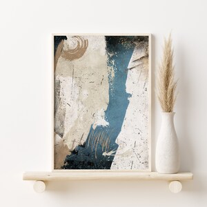 May include: Abstract art print with blue, white, and gold paint strokes on a textured canvas. The print is framed in a thin, light wood frame and is displayed on a white shelf.