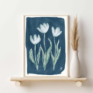 May include: A framed art print featuring three white tulip flowers with green stems and leaves against a dark blue background. The artwork is in a light wooden frame and is signed in the bottom right corner. The print is displayed on a wooden shelf.