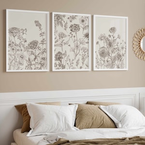 May include: Three framed botanical prints with detailed line drawings of various flowers and plants. The prints are in white frames and hang above a bed with white and brown pillows. A round, woven mirror and a rattan screen are also visible.