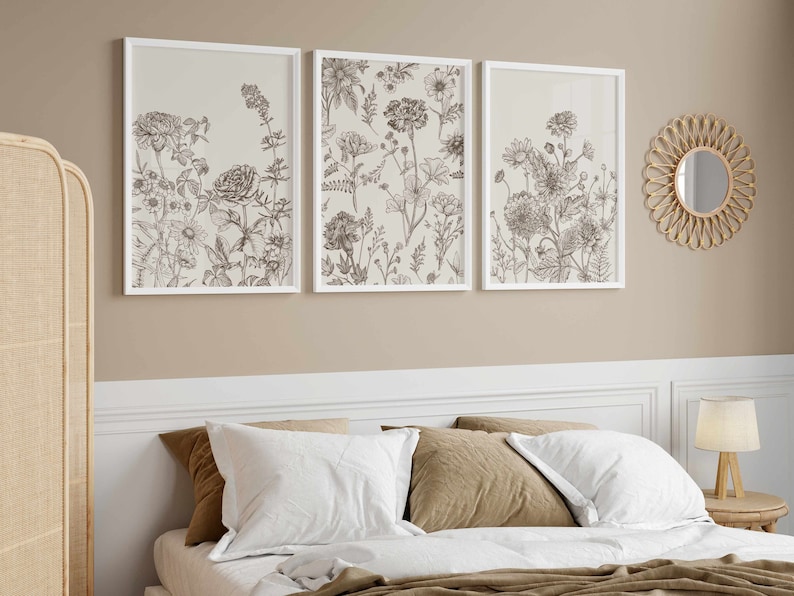 May include: Three framed botanical prints with detailed line drawings of various flowers and plants. The prints are in white frames and hang above a bed with white and brown pillows. A round, woven mirror and a rattan screen are also visible.