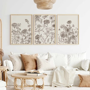 May include: Three framed botanical art prints featuring detailed line drawings of various flowers in shades of brown and beige. The prints are displayed above a white sofa with neutral-toned pillows and a wooden coffee table.