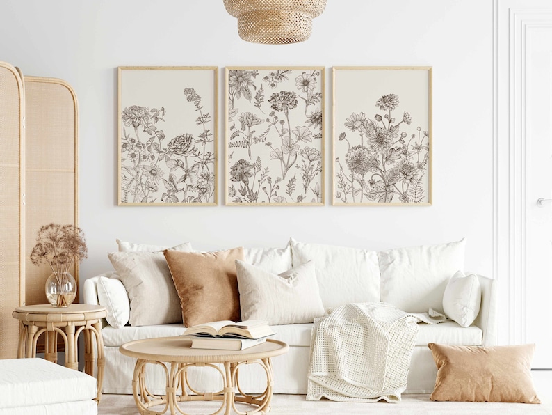 May include: Three framed botanical art prints featuring detailed line drawings of various flowers in shades of brown and beige. The prints are displayed above a white sofa with neutral-toned pillows and a wooden coffee table.