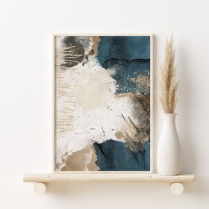 May include: Abstract art print with blue, white, and gold paint splatters on a canvas. The print is framed in a light wood frame and is displayed on a shelf.