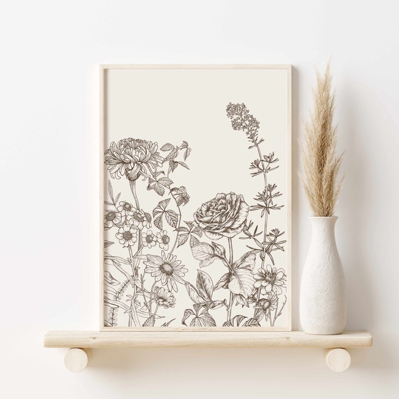May include: A framed botanical print featuring detailed line art of various flowers and foliage in shades of brown and beige. The artwork is displayed on a light wooden shelf, with a white vase holding dried grass to the right.