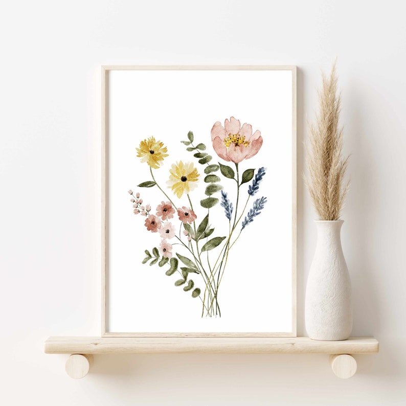 May include: Watercolor floral print featuring a bouquet of flowers in shades of yellow, pink, and blue. The artwork is framed in a light wood frame and displayed on a wooden shelf. A vase with dried grass is to the right.