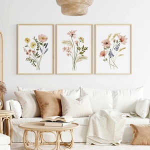 May include: Three framed floral watercolor prints. Each print features a different bouquet of flowers in shades of pink, yellow, and blue, with green foliage. The frames are a light wood color, and the prints are displayed above a white sofa.