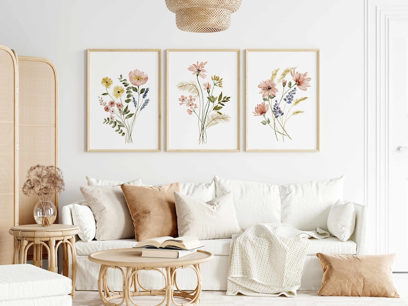 May include: Three framed floral watercolor prints. Each print features a different bouquet of flowers in shades of pink, yellow, and blue, with green foliage. The frames are a light wood color, and the prints are displayed above a white sofa.