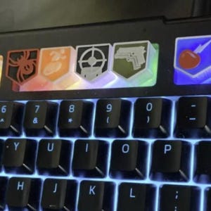 Peut inclure: Un clavier d'ordinateur noir avec une rangée de touches personnalisées rétroéclairées au-dessus des touches numériques. Les touches affichent diverses icônes colorées, dont une pierre tombale avec RIP, une araignée, une cible et un pistolet.
