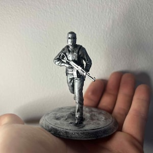 May include: A silver figurine depicting a soldier running and holding a rifle. The figure wears a mask and tactical vest, mounted on a circular base. The figurine is held in a hand, with a plain background.