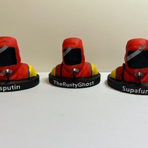 Puede incluir: Tres figuras impresas en 3D de un personaje que lleva un mono rojo con capucha, una máscara facial negra y una franja de cinta plateada. Las figuras están sobre bases negras con los nombres "Rasputin", "TheRustyGhost" y "Supafunki" impresos en ellas.