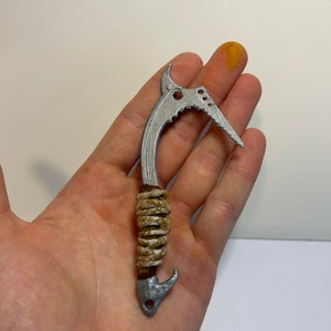 Peut inclure: Un crochet en métal argenté avec une poignée en corde. Le crochet a une bordure dentelée et une pointe pointue. La poignée en corde est enveloppée de ficelle brune.