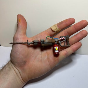 May include: A miniature, hand-painted, rusty-looking drill with a red and yellow warning label. The drill has a long, thin, metal drill bit and a brown wooden handle.