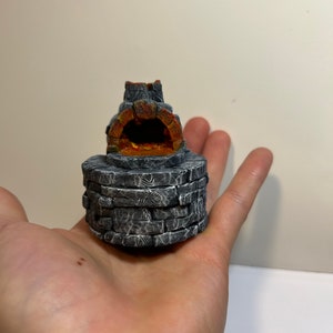 May include: A small, grey stone oven with a rounded opening and a textured surface. The oven is lit with orange flames inside.