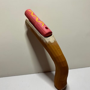 May include: A wooden toy gun shaped like a hot dog with a pink bun and yellow mustard. The gun is painted brown and has a textured surface.
