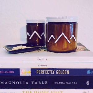 May include: Two amber glass candle jars with white lids and white mountain range designs. The candles are stacked on top of a stack of books with titles like "Kinfolk", "Perfectly Golden", "Magnolia Table", "The Home Edit", and "The Kinfolk Table".