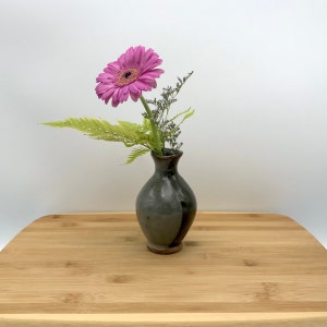 May include: A single pink gerbera daisy with green fern leaves in a small, dark gray ceramic vase. The vase has a crackled glaze and a dark gray stripe down the side. The vase is sitting on a light brown wooden surface.