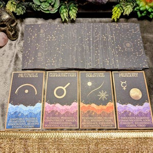 May include: A deck of tarot cards with a celestial theme. The cards are black with gold star patterns and feature words like "Mutable" and "Conjunction." The bottom of the cards have descriptions. The deck is on a dark surface.