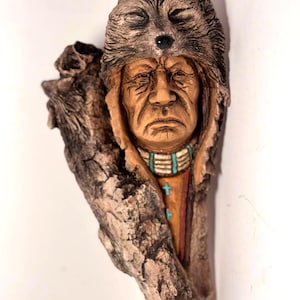 May include: A carved wooden sculpture of a Native American man wearing a wolf headdress. The sculpture is mounted on a piece of driftwood. The man's face is detailed, and he wears a beaded necklace and a colorful shirt. The wood has a natural, textured appearance.