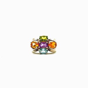 May include: A silver ring featuring a cluster of five oval-cut gemstones arranged in a cross pattern. The center stone is purple, surrounded by a lime green stone at the top, a light blue stone at the bottom, and orange stones on each side.