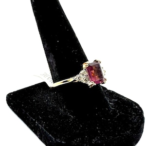 May include: A gold ring with a rectangular, reddish-purple gemstone and small, clear accent stones. The ring is displayed on a black velvet ring holder. The ring's band is thin and appears to be gold.
