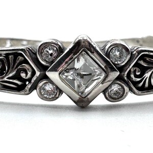 May include: Close-up of a silver ring with a square, clear gemstone in the center, surrounded by smaller, round clear gemstones. The ring has an ornate, scrollwork design on the sides.