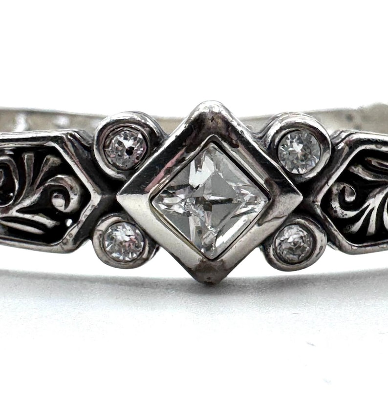 May include: Close-up of a silver ring with a square, clear gemstone in the center, surrounded by smaller, round clear gemstones. The ring has an ornate, scrollwork design on the sides.