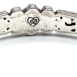 May include: Close-up of a silver-toned bracelet. The bracelet features a decorative design with a heart-shaped logo. The metal has a textured appearance with dark accents. The bracelet's design includes a series of raised, ridged elements.