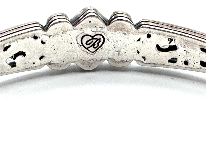 May include: Close-up of a silver-toned bracelet. The bracelet features a decorative design with a heart-shaped logo. The metal has a textured appearance with dark accents. The bracelet's design includes a series of raised, ridged elements.