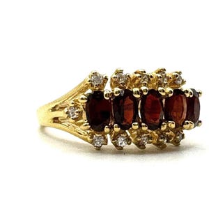 May include: A gold ring featuring five oval-cut, deep red gemstones, accented with small, round, clear gemstones. The ring has a detailed, ornate design and is set against a white background.