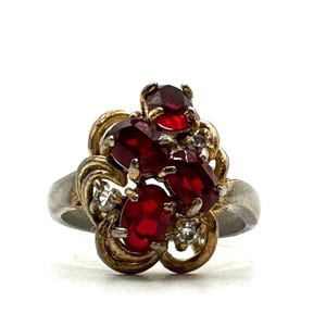 May include: Vintage ring featuring a cluster of oval-cut, deep red gemstones, accented with smaller clear stones and gold-toned filigree detailing. The band is silver-toned. A classic piece of jewelry.