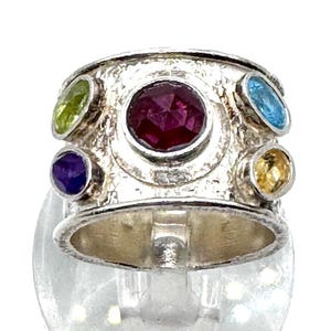 May include: A silver ring with a textured band, featuring a large, round, deep red gemstone at the center, surrounded by smaller, colorful gemstones in green, blue, yellow, and purple. The ring is displayed on a clear stand.