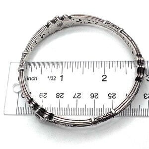 May include: Silver-toned bangle bracelet with intricate etched designs and decorative accents. The bracelet is measured with a ruler, showing a diameter of approximately 2.75 inches. The jewelry piece has a vintage or antique aesthetic.