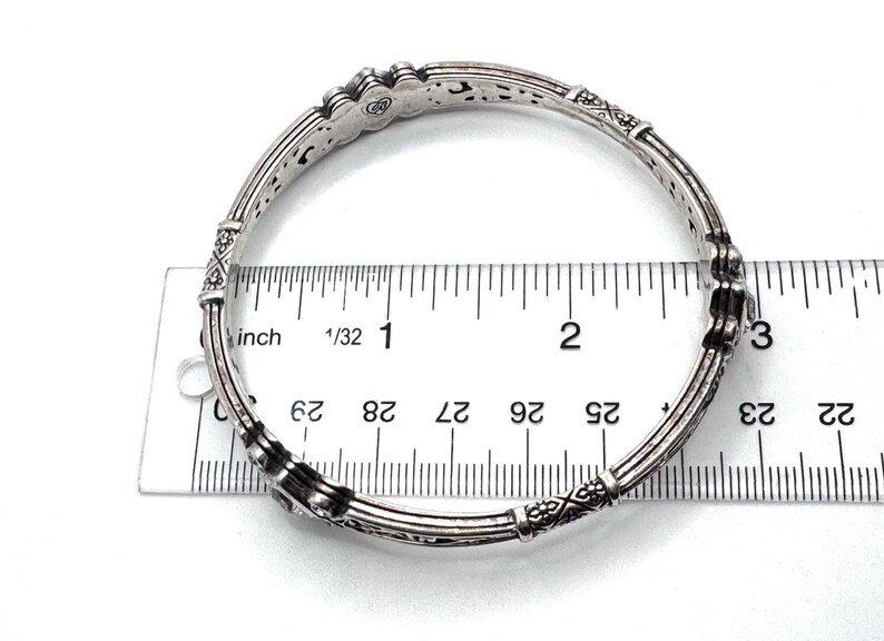 May include: Silver-toned bangle bracelet with intricate etched designs and decorative accents. The bracelet is measured with a ruler, showing a diameter of approximately 2.75 inches. The jewelry piece has a vintage or antique aesthetic.