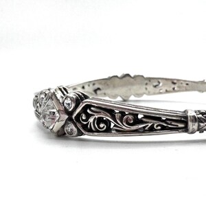 May include: A silver-toned bracelet with an ornate design. The bracelet features a central diamond-shaped stone and smaller round stones. Intricate scrollwork and filigree patterns adorn the band, creating a vintage or antique aesthetic.