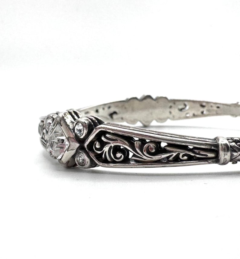 May include: A silver-toned bracelet with an ornate design. The bracelet features a central diamond-shaped stone and smaller round stones. Intricate scrollwork and filigree patterns adorn the band, creating a vintage or antique aesthetic.