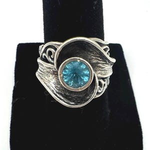 May include: A silver ring with a large, round, light blue gemstone at the center. The ring has a textured, swirling design around the gem. The ring is displayed on a black surface.