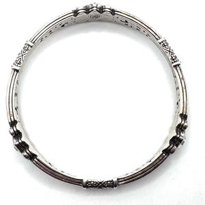 May include: A silver-toned bangle bracelet with intricate detailing. The bracelet features decorative floral and geometric patterns, with a series of vertical bars and connecting segments. The design is symmetrical, with a repeating pattern around the entire circumference.
