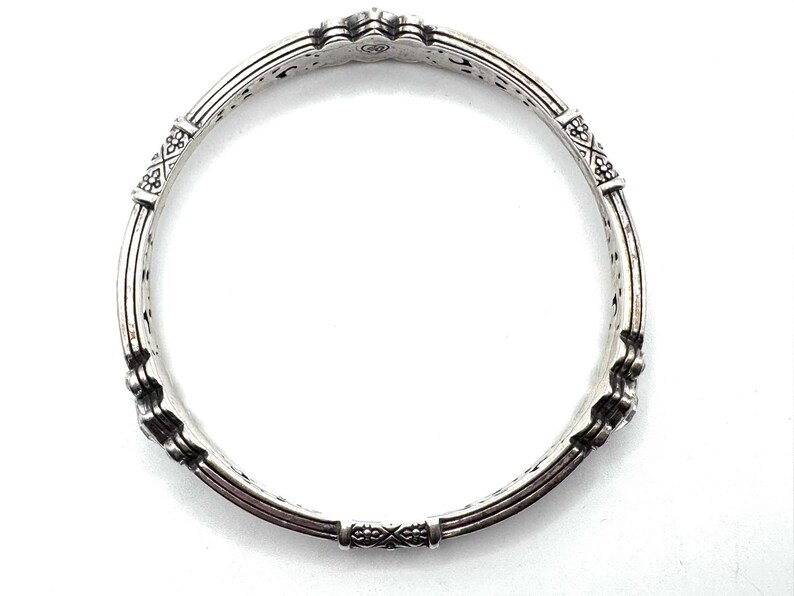 May include: A silver-toned bangle bracelet with intricate detailing. The bracelet features decorative floral and geometric patterns, with a series of vertical bars and connecting segments. The design is symmetrical, with a repeating pattern around the entire circumference.