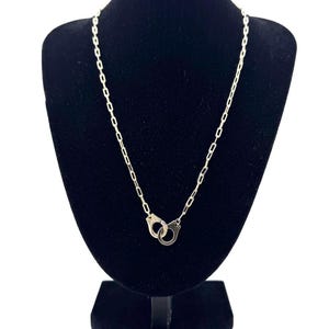 May include: A silver-toned metal chain necklace featuring a small, detailed handcuff charm as the central pendant. The necklace consists of elongated rectangular links and is displayed on a black velvet-textured neck form for clear visibility.