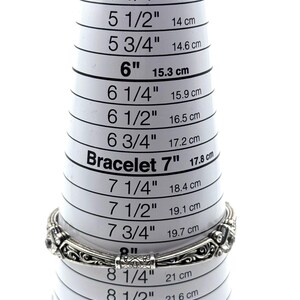 May include: A silver bracelet with an ornate design is placed on a white EZ Bracelet Mini sizer. The sizer shows measurements in inches and centimeters, with the bracelet fitting at approximately 7 inches (17.8 cm).